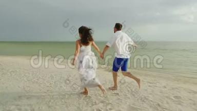 青年男女牵着手，赤脚沿着沙滩奔跑.. 节日和蜜月快乐。 4K慢动作视频